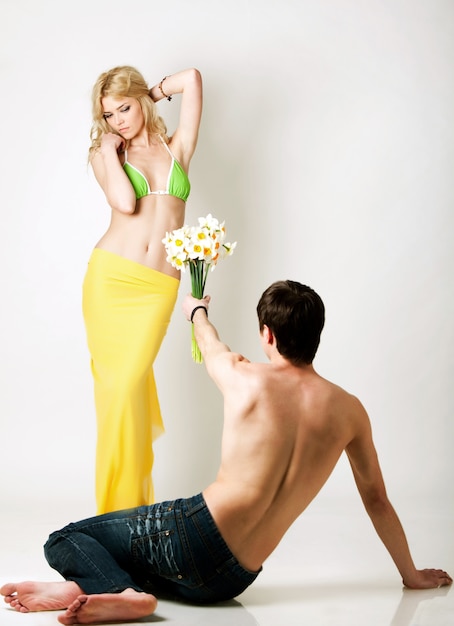 Young man presenting flowers to blond beautiful woman in green bikini and yellow pareo over white background in photo studio. Beauty and fashion lifestyle concept