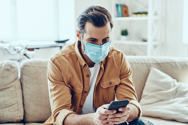 Young man in protective face mask using smart phone while working from home