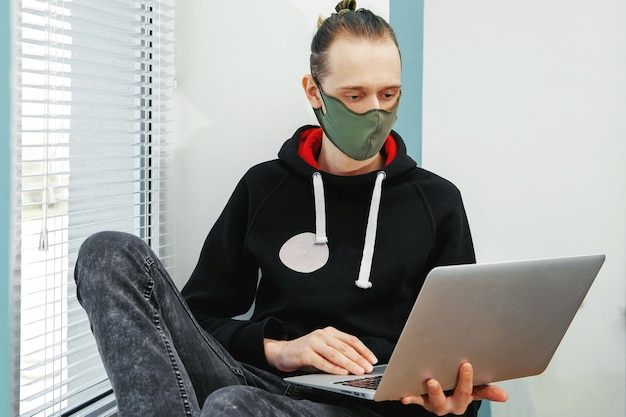 A young man in a protective mask sits at a window and works on a computer. Coronavirus, work from home in isolation.
