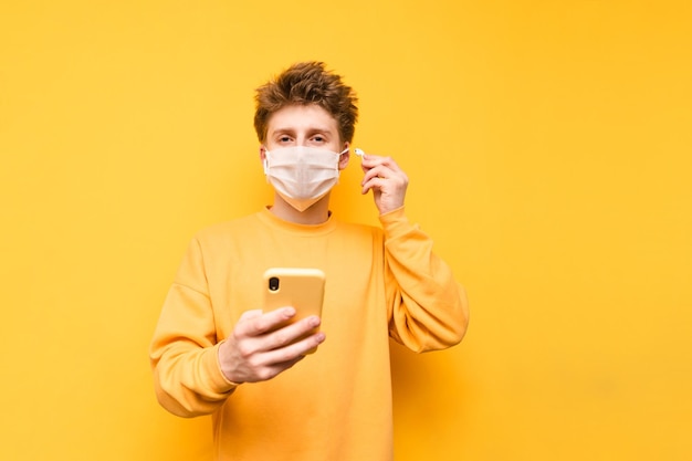 A young man in a protective mask stands on a yellow background with a smartphone in his hand