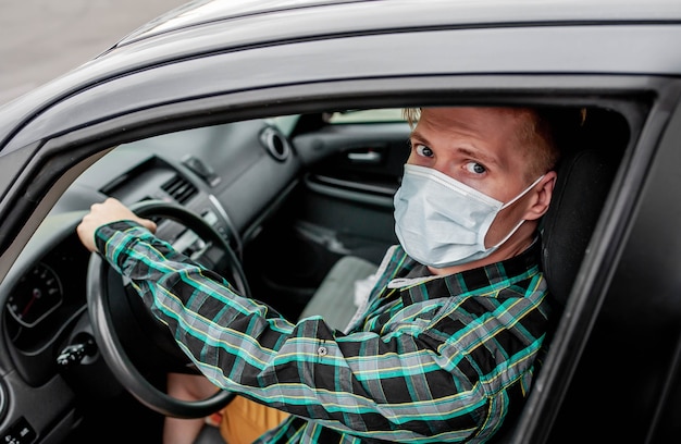 A young man in a protective sterile medical mask is sitting behind the wheel of the car. COVID-19.