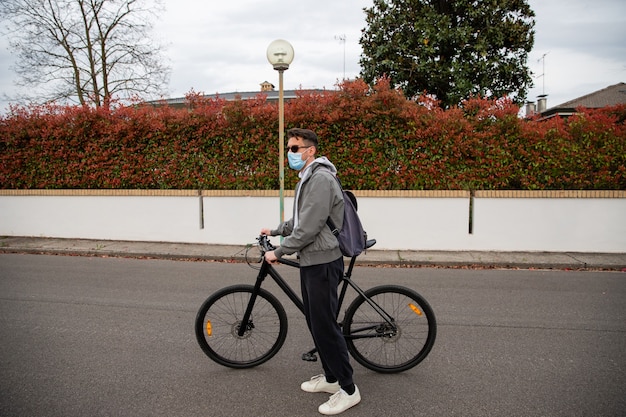 若い男が手で自転車を押し、通りに立ってサージカルマスクを着用します