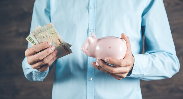 Young man putting banknote into piggy bank on color background