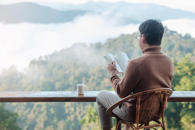 窓際で本を読み、朝日が昇る田舎のホームステイで山の景色を眺める青年。 SoloTravel、旅、旅行、リラックスのコンセプト