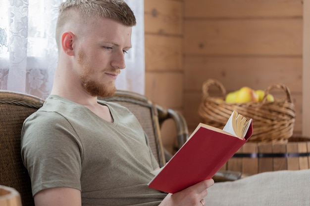 Young man reading book with red cover on wicker bench in countryside wooden house against background of basket of yellow apples