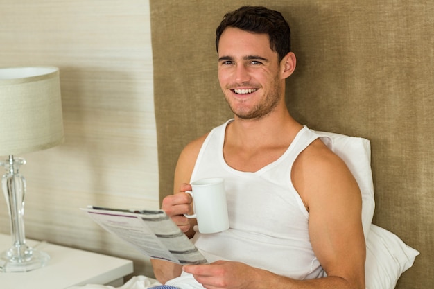 Young man reading newspaper while holding a cup of tea in bedroom