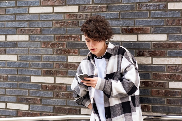 젊은 남자가 벽돌 벽에 맞춰 전화로 무언가를 읽습니다.