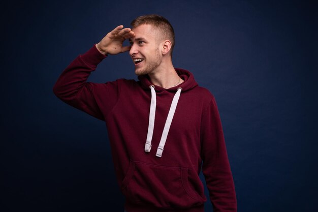 A young man in a red hoodie and jeans poses on a blue background, puts his hand to his face and looks into the distance