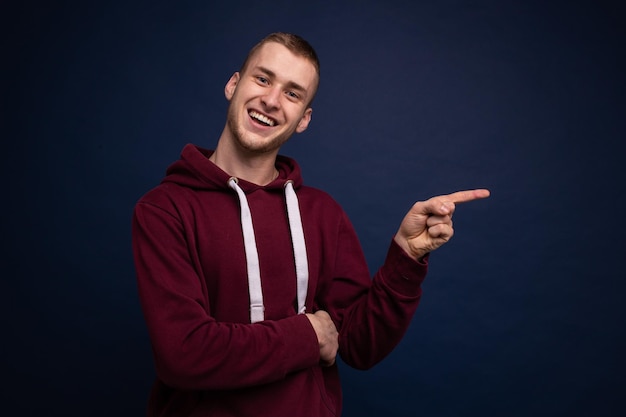 Young man in red hoodie and jeans posing on a blue background, smiling and showing a finger to the side