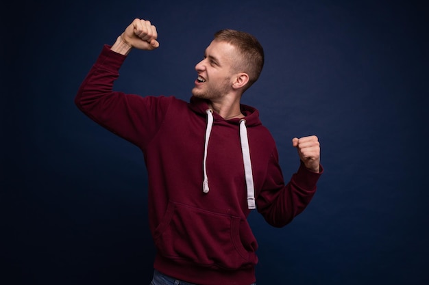 Young man in red hoodie and jeans posing on blue background smiling showing victory