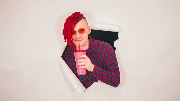 Young man in red plaid shirt drinking through soft drink straw in hole of white background Bright guy enjoying nonalcoholic beverage