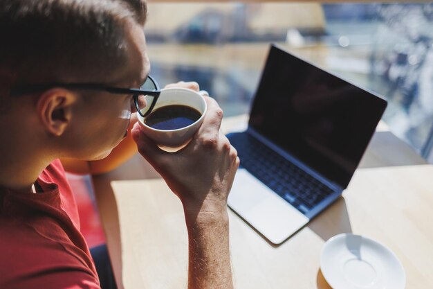 A young man in a red tshirt glasses for vision correction on\
his face he stands at the window with a cup of coffee and looks at\
the city