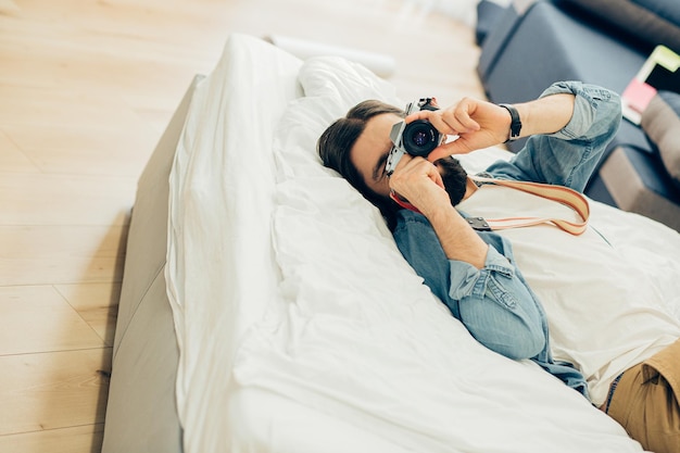 Young man relaxing on the bed at home and taking photos on his camera