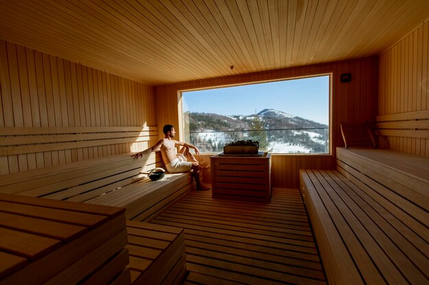 Young man relaxing in the sauna and watching winter forest through the window