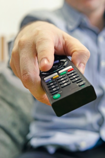 Young man relaxing on sofa and holding TV remote control. the TV remote is in the foreground. Switching channels. vertical photo