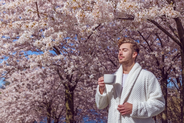 Young man relaxing in the spring blooming garden and drinking coffee from the cup