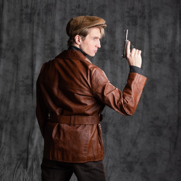 Молодой человек ретро секретный агент парень в коричневой кожаной куртке и плоской кепке с пистолетом в руке