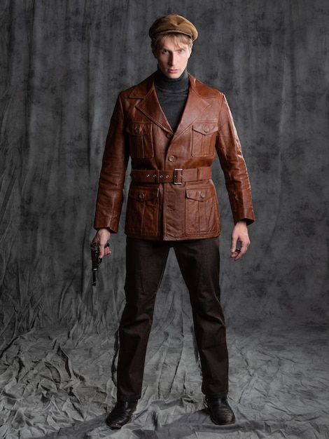 Молодой человек ретро секретный агент парень в коричневой кожаной куртке и плоской кепке с пистолетом в руке