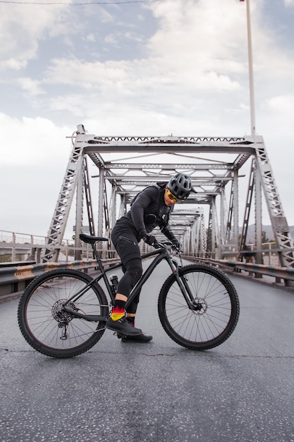 Young man riding a bicycle wearing cycling equipment in an\
urban steel bridge