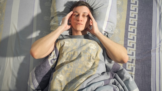 Young man rubbing temples while lying on comfortable bed and feeling unwell