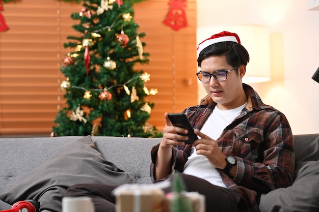 Young man in Santa hat using smart phone while sitting on couch near the Christmas tree at home.