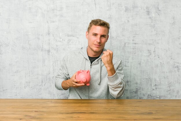 Young man saving money with a piggy bank showing fist to camera, aggressive facial expression.