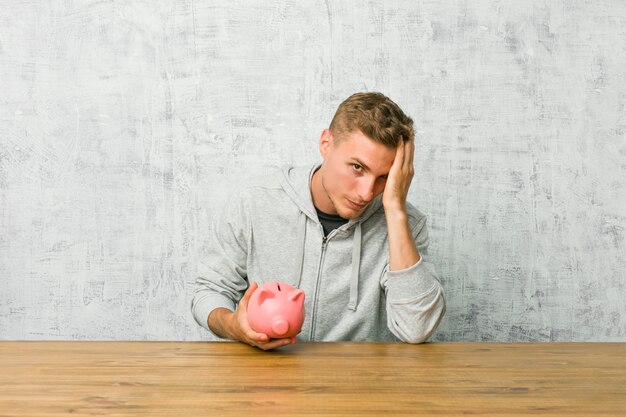 Young man saving money with a piggy bank tired and very sleepy keeping hand on head.