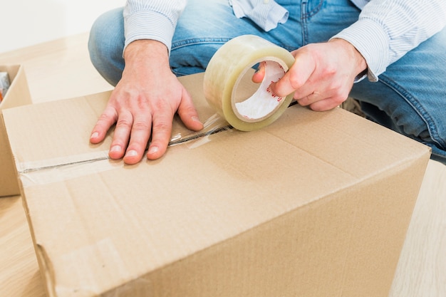 Фото Молодой человек запечатывает картонную коробку с лентой большой для перемещения