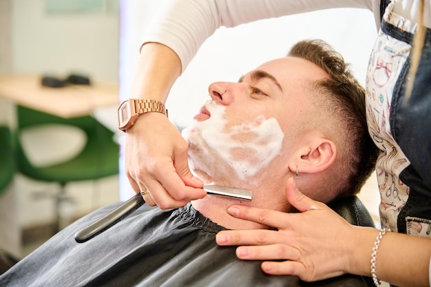 Young man shaving with straight edge razor by hairdresser in barbershop