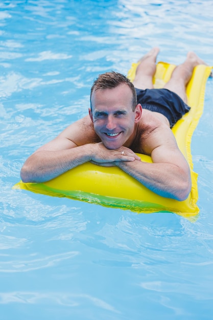 반바지 차림의 한 청년이 카메라를 보며 웃고 있는 반짝이는 푸른 수영장에 있는 큰 고리에 떠 있는 워터파크를 즐깁니다. 여름 방학
