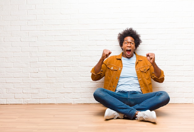 Young man shouting aggressively with an angry expression or with fists clenched celebrating success sitting on the floor at home