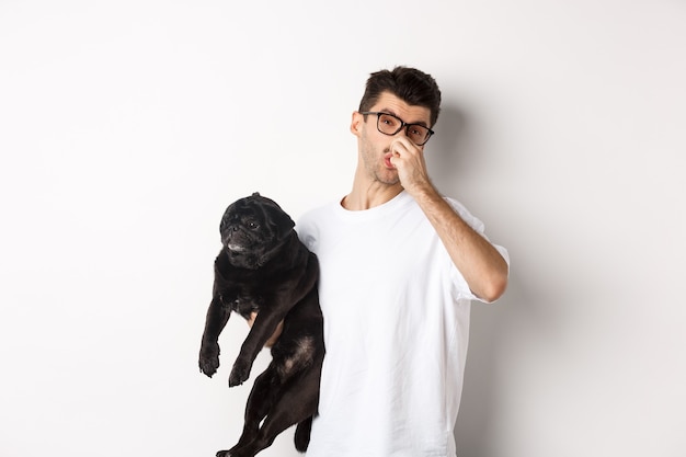 Young man shut nose as holding pug, disgusted with bad smell fart of animal, standing over white