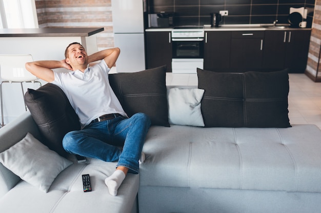 Young man sit on couch and laughing out loud. Loook up and hold hands under head. Relax, rest and enjoy. Alone in room. Daylight.