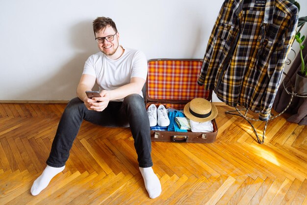 Young man sit on floor checking list on mobile valise packing for triptravel concept copy space