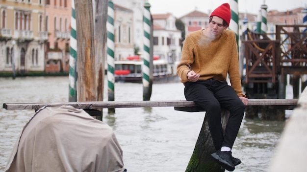 A young man siting on the bench uper the waterfront and and brutally smokes cigarettes