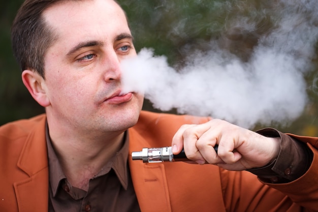 A young man sits on a bench and smokes an electronic cigarette