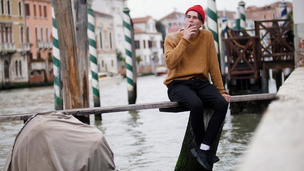 A young man sits on the bench above the water and smokes