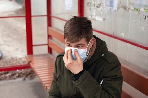 Young man sits at bus station in disposable medical mask close-up.