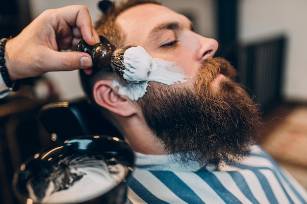 A young man sits in a chair while a barber styles his beard.