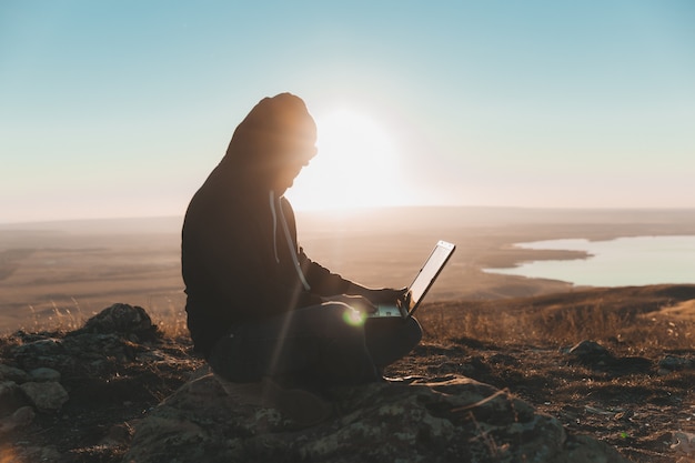 Un giovane si siede su una collina e usa un laptop. nomadi digitali.