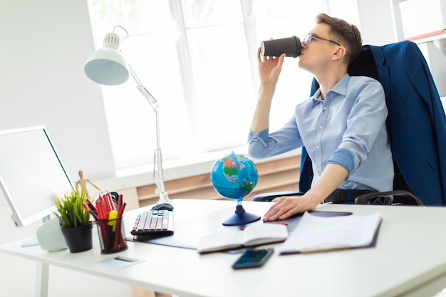 A young man sits in the office at a computer desk, holds a globe with his hand and drinks coffee.