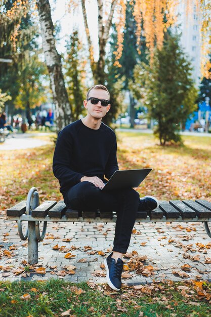 A young man sits in the park on a bench and works on a laptop a\
warm autumn day remote work