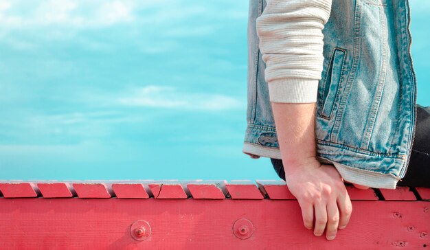 A young man sits turned sideways on a red wooden pier against a bright blue sky. Summer lonely man travel banner with space for text