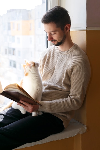 若い男が窓辺に座って本を読みます。男の隣に赤い斑点のある白い猫が座っています。男性はベージュのセーターを着ています。