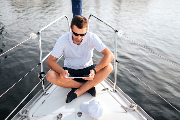 Photo young man sits with his legs crossed and hold tablet in hands. he is calm and concentrated. screen is black. guy sits on yacht bow. he wears sunglasses.