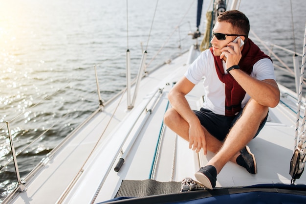 Young man sits on yacht board and look to left. He talks on phone. Guy sits with legs crossed. He wears sunglasses. Young man is busy.