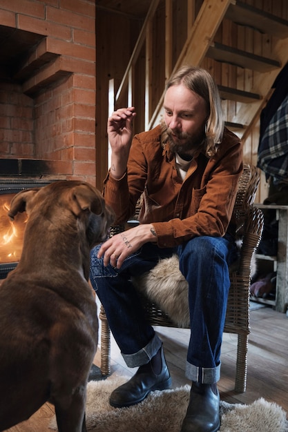 Young man sitting on armchair near the fireplace and training his dog during leisure time at home