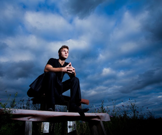 들판 한가운데 벤치에 앉아 있는 젊은 남자. 어두운 푸른 하늘 구름 배경