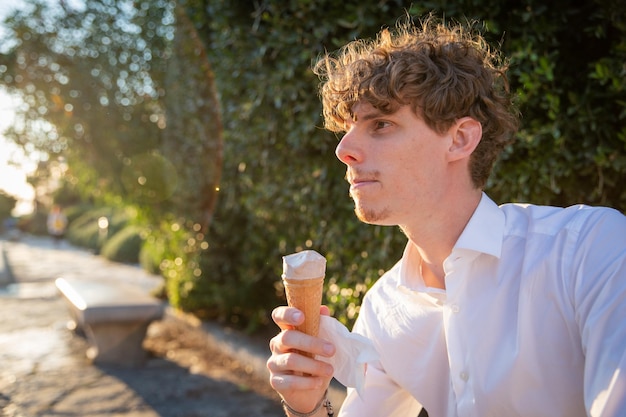 公共の公園のベンチに座っている若い男は、左側の晴れた日のコピー スペースでアイスクリームを楽しんでいます