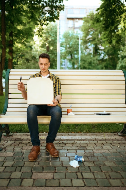Молодой человек сидит на скамейке среди мусора в парке, занимается волонтерством